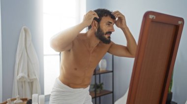 Handsome young hispanic man with a beard checking his hair in the mirror at an indoor spa beauty salon. clipart