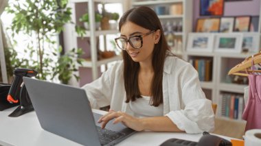 Dizüstü bilgisayar mağazasında çalışan genç bir kadın. Arka planda kitaplar, dekorasyonlar ve bitkiler var.