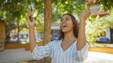 Şehir parkında kutlama yapan bir kadın, elinde bir telefon, parlak güneş ışığında bir ağacın altında gülümsüyor.