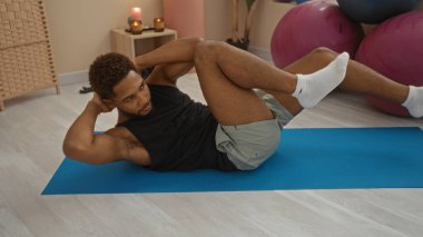 Spor salonundaki mavi bir paspasın üzerinde egzersiz yapan genç bir adam kararlı bir ifadeyle çekirdek gücünü gösteriyor.