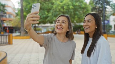Anne ve kızı bir şehir parkında selfie çekiyorlar, yeşillik ve şehir binalarıyla çevrili mutlu bir şekilde gülümsüyorlar, kadınlar arasındaki sevgi ve dostluğu yakalıyorlar.