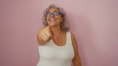 Güzel, yaşlı, beyaz saçlı, pembe arka planda izole olmuş, gülümseyen ve işaret eden İspanyol bir kadın.