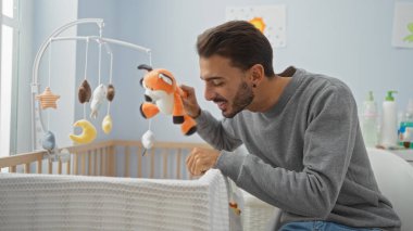 Genç adam, beşikte beşiği ve yumuşak ışığı olan rahat yatak odasında bebeğe oyuncak gösteriyor. Sevimli ve neşeli bir ev ortamı öneriyor..