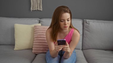 Pembe bluzlu genç bir kadın gri bir koltukta oturmuş dekoratif yastıklarla dolu modern bir oturma odasında telefonuna bakıyor.