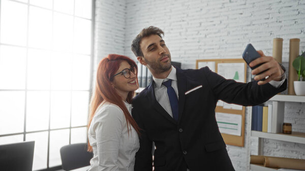 Man and woman taking selfie in modern office space smiling wearing formal attire with shelves and desk showing a professional indoor setting indicating business partnership.