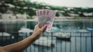 Elinde liman ve arka planda tekneler olan Türk lira banknotları, açık deniz kenarı finans temasını gözler önüne seriyor..
