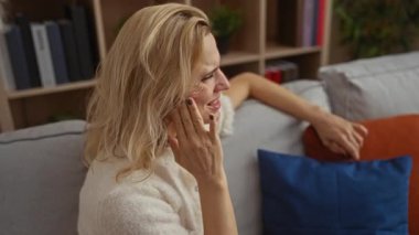 Dişleri ağrıyan bir kadın, rahat bir oturma odasında, rafları ve yastıkları olan, rahatsız edici ve endişeli bir şekilde otururken, ev ortamını yansıtan ev unsurlarıyla çevrelenmiş..