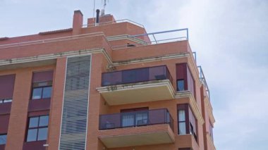 Mavi gökyüzünün altında balkonları ve kırmızı tuğlalı cephesi olan modern apartman binası, huzurlu bir mahalle ortamında kentsel mimari ve çağdaş tasarım sergiliyor..