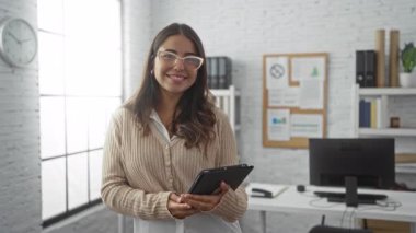 Modern ofiste elinde tabletle gülümseyen bir kadın. Parlak iş yerinde gözlük takıyor. Masa rafları ve saat beyaz tuğla duvara dayanıyor..