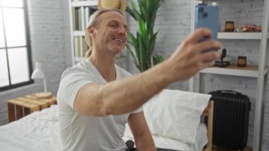 Beyaz çarşaflı ve şık dekorlu modern yatak odasında selfie çeken sarışın beyaz bir adam bir neşe ve rahatlama anı yakalıyor..