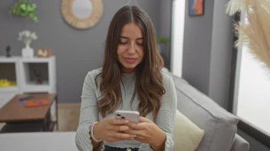 Kadın rahat oturma odasında akıllı telefon kullanırken gülümsüyor, mutluluğu ve modern teknolojiyle bağlantıyı sergiliyor, rahat bir şekilde koltukta oturuyor..