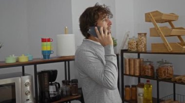 Ofis mutfağında telefonla konuşan genç adam çeşitli renkli mutfak aletleri ve ahşap raflarla çevrili, gündelik ve modern bir çalışma alanı ortamı yaratıyor..