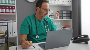 Bir hastane ofisinde dizüstü bilgisayar ve telefonla çalışan, notlar yazan, yeşil önlük giyen, tıbbi malzeme ve dosyalarla çevrili görevlere odaklanmış bir doktor..