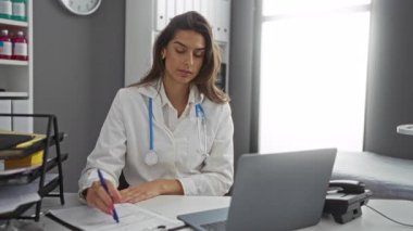 Klinikte dizüstü bilgisayar ve belgelerle oturan kadın doktor, parlak bir odada çalışmaya odaklanmış, profesyonellik ve kendini adamışlık sergiliyor..