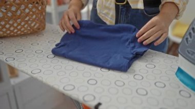 Çamaşır odasındaki ütü masasında giysileri katlayan kadın ev işlerini ve organizasyonu görünür ellerle, kapalı alanlarda sergiliyor..