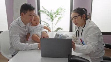 Babası bebeği klinikte tutarken dizüstü bilgisayar kullanan, hastane ortamında aile sevgisini ve bakımını gösteren kadın bir çocuk doktoruyla konuşuyor..