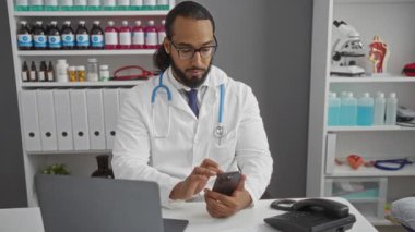 Hastane ofisinde akıllı telefon kullanan bir doktor. Masasında dizüstü bilgisayar ve telefonla. Etrafı tıbbi malzeme ve modern sağlık hizmetlerini yansıtan ekipmanlarla çevrili..