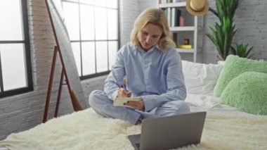 Konforlu yatak odasında dizüstü bilgisayarı olan, pijama giyen, bacak bacak üstüne atmış, doğal ışıkta yıkanmış, yastıklarla çevrili, huzurlu bir ev dekorasyonu sergileyen bir kadın..