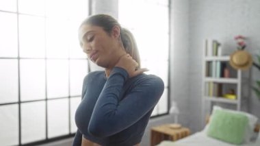 Modern ev ortamında boynu ağrıyan, gün ışığında mavi bluz ve at kuyruğu saçı takan bir kadın yatak odasında..