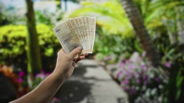 Danimarka Kronu 'nu elinde tutan adam canlı bir bahçe parkında, yemyeşil ve renkli çiçeklerle birlikte..