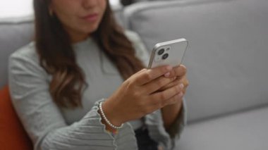 Ev ortamında esmer saçlı, akıllı telefon kullanan bir kadın rahat bir oturma odasında odak ve teknoloji sergiliyor..