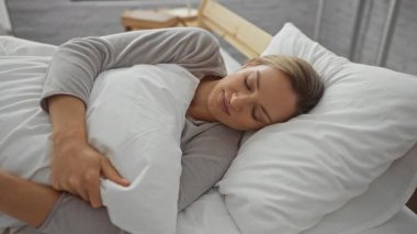Yatakta huzur içinde uyuyan bir kadın rahat bir yatak odasında yastığa sarılıyor sakin bir ifadeyle ve sakin bir atmosferle rahatlama ve huzuru gösteriyor..
