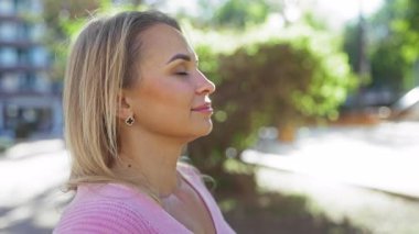 Güneşli şehir parkında huzurlu bir anın tadını çıkaran bir kadın, gözleri kapalı, pembe bir kazak giyiyor, yeşillikle çevrili, dış mekan sakinliği ve rahatlığı somutlaştırıyor..