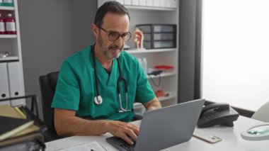 Modern bir klinikte dizüstü bilgisayarda yazan yeşil önlüklü bir doktor. Boynunda steteskop ve arka planında tıbbi malzemelerle dolu raflar var..