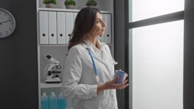 Klinikteki kadın elinde steteskopla elinde kahve bardağıyla pencereden dışarı bakıyor. Rafın yanındaki parlak geniş pencereden gözetliyor..