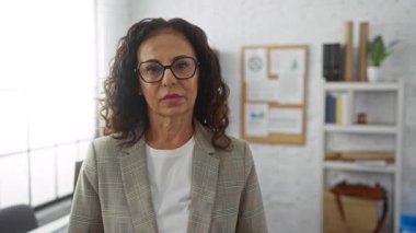 Kıvırcık saçlı olgun bir kadın ofiste gözlüklü, spor ceketli, profesyonelliğini ve özgüvenini aktaran arka planda kitap ve kağıtlarla duruyor..