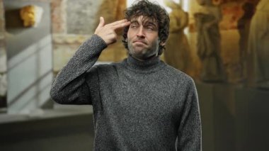 İçerideki genç adam rahat bir ifadeyle el hareketi yapıyor gri kazak giyiyor loş bir kilise ortamında duruyor ve arka planda sanatsal heykeller var..