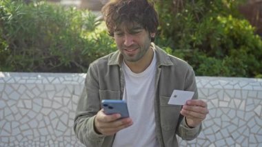Genç adam, elinde akıllı telefon ve kredi kartıyla şehir parkında gülümsüyor. Açık hava dijital ödeme etkileşimini yansıtıyor..