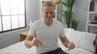 Sarışın, orta yaşlı bir adam, geniş pencereli ve yeşil bitkilerli, parlak, modern bir yatak odasında neşeyle dans ediyor. Rahat, pozitif bir ev atmosferi sergiliyor..