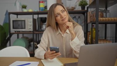 Ev telefonuyla düşünen bir kadın. Etrafı dizüstü bilgisayar, not defteri, mikrodalga fırın ve raflarla çevrili. Modern iş yaşam tarzını resmediyor..