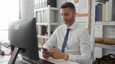 Modern ofis ortamında telefon kullanan bir adam, etrafında bilgisayar ve belgelerle, odak, neşe ve başarı ifadelerini gösteriyor..
