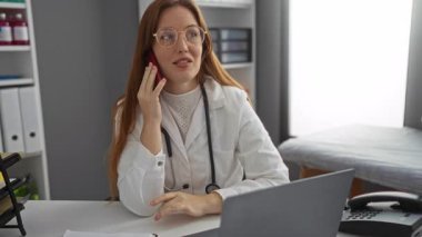 Klinikte telefonla konuşan, iş yerinde steteskop takan beyaz önlüklü bir kadın, modern iç tasarımı olan profesyonel bir sağlık ortamını işaret ediyor..
