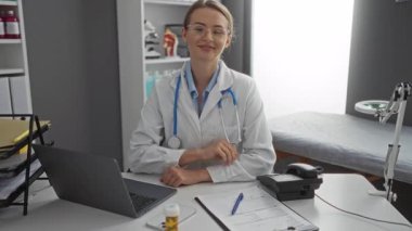 Klinikte gülümseyen, gözlüklü, steteskop laptoplu ve masasında profesyonel bir sağlık sigortası olduğunu gösteren belgeler olan bir kadın..