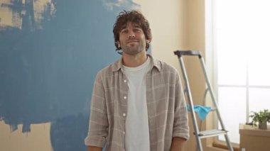 Genç İspanyol adam evin içini mavi duvarlı merdiven ile boyuyor. Evin içinde gülümsüyor.