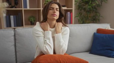 Oturma odasında kanepede oturan, boynu ağrıyan bir kadın, rahatsızlık ifadesi gösteren, modern bir dairede kitap ve bitkilerle çevrili, rahat bir kazak giyen bir kadın..