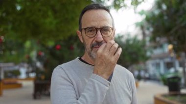Olgun İspanyol bir adam güneşli bir şehir parkında jest yapıyor. Yeşillik ve şehir manzarası var. Kendine güveni ve düşünceli..
