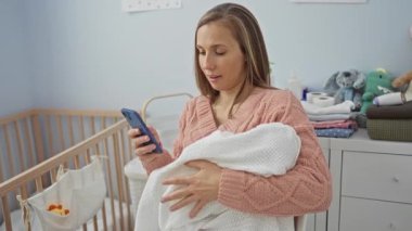 Bebeği kucağında tutan ve rahat yatak odasında beşiği ve oyuncakları olan akıllı telefon kullanan, ev kadınlığını sergileyen ve huzurlu bir ev ortamında birden fazla iş yapan bir kadın..