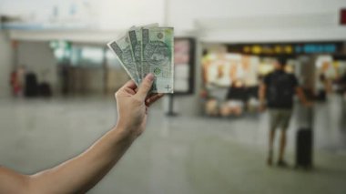 Arka planda bulanık gezginler ve uçuş tabelaları olan kapalı bir havaalanında, seyahat masraflarını ve döviz kurunu vurgulayan, Polonya zloti banknotları tutan adam..
