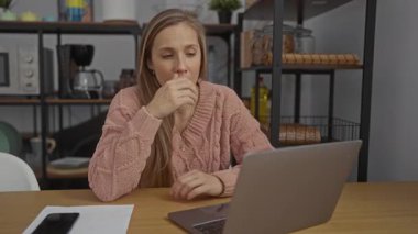 Pembe kazaklı kadın, ofis ortamında yorgun bir ifadeyle dizüstü bilgisayara bakıyor. Raflar ve mutfak eşyalarıyla çevrili. Yorgunluk ve iş stresi hissi veriyor..