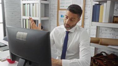 Beyaz gömlekli ve mavi kravatlı kendinden emin bir adam, modern ofisteki bilgisayar başında profesyonellik ve verimliliği gösteren belgeler ve kitaplarla çevrili çalışmayı tartışıyor.
