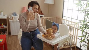 Yatak odasında, bebek sandalyesinde oyuncaklarla çevrili bir şekilde telefonla konuşan, ahşap zemin ve doğal ışıkla döşenmiş sıcak ev ortamında sevgi ve bağlarını ifade eden bir kadın..