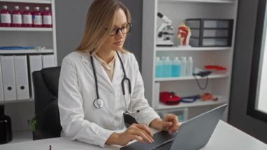 Klinikteki kadın doktor dizüstü bilgisayarda çalışıyor. Profesyonel tıbbi ortamda steteskop takıyor. Rafları ve ekipmanları içeride.