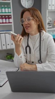 Klinikteki kadın doktor, gözlüklü, steteskop takan, bilgisayarlı, kapalı alanda profesyonel raflar kuran, akıllı telefonlardan konuşuyor.