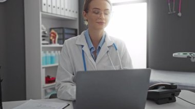Klinikteki beyaz önlüklü, gözlüklü, profesyonel tıbbi çalışma alanında dizüstü bilgisayara gülümseyen, steteskoplu ve raflarda organize edilmiş belgeleri olan bir kadın..