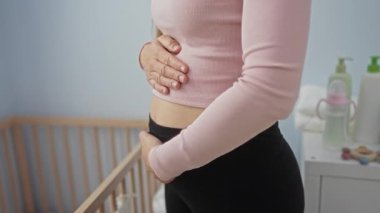 Genç bir kadın yatak odasında karnına hafifçe dokunuyor. İki eli bebek beşiğinin yanında duruyor. Annelik ve bakım temalarını ima ediyor..