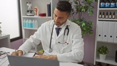 Steteskoplu beyaz önlüklü bir klinikte çalışan, telefonlara cevap veren ve notlar alan, profesyonel bir ofiste tıbbi ekipman ve bitkilerle çevrili bir doktor..